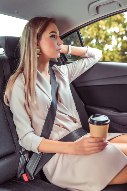 Business woman taxi car, summer city, resting after work and looking out window, holding cup coffee tea, having snack for breakfast lunch, waiting for trip. Casual makeup pink formal suit. - Photo, Image