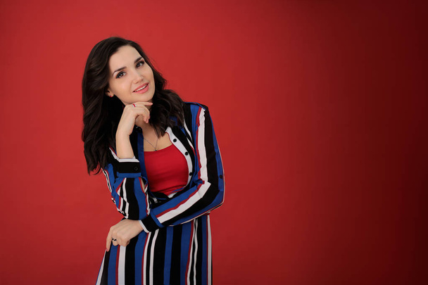 portrait of a beautiful girl with dark hair in a striped dress on a red background in a good positive mood, a young woman free space for notes - Foto, imagen