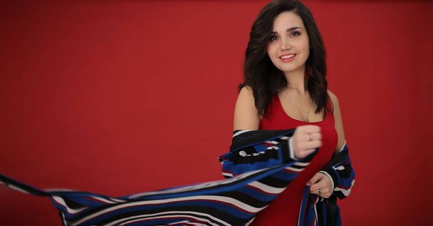portrait of a beautiful girl with dark hair in a striped dress on a red background in a good positive mood, a young woman free space for notes - Foto, imagen