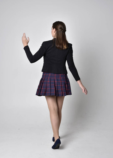 full length portrait of a pretty brunette girl wearing a school uniform of black jacket and plaid skirt. Standing pose, with back to the camera, isolated against a studio background. - Foto, afbeelding