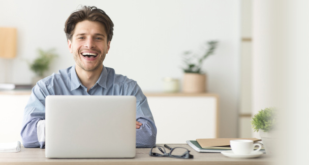 Щасливий працівник посміхається на камеру під час роботи на ноутбуці в офісі
 - Фото, зображення