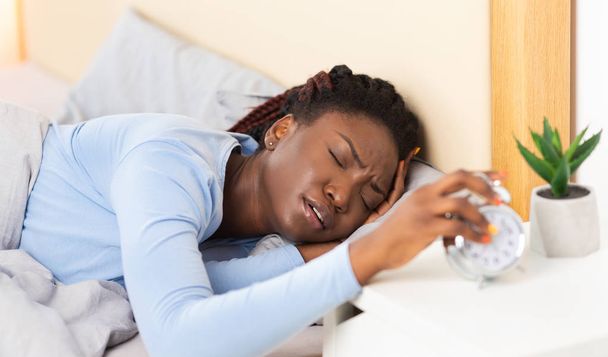 schläfriges schwarzes Mädchen schaltet Wecker aus, der im Bett liegt - Foto, Bild