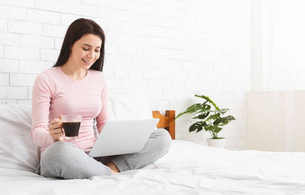 Millennial girl sitting on bed, working on laptop with coffee - Fotó, kép