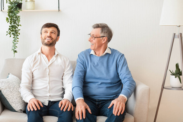 fröhlicher erwachsener Sohn und älterer Vater sprechen zu Hause auf dem Sofa sitzend - Foto, Bild