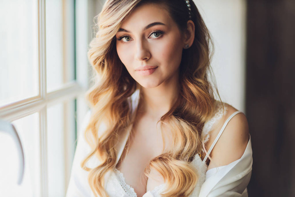 Portrait of a beautiful blonde bride in the interior. Morning young bride in a bathrobe. Wedding photography. Smiling and cute girl stands. - Foto, imagen
