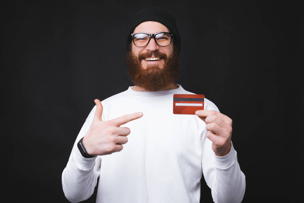 Smiling bearded man wearing eyeglasses and pointing at red credit card - Zdjęcie, obraz