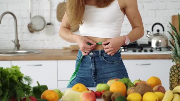 Girl measures her waist in the kitchen near the table with vegetables and fruits. - Πλάνα, βίντεο