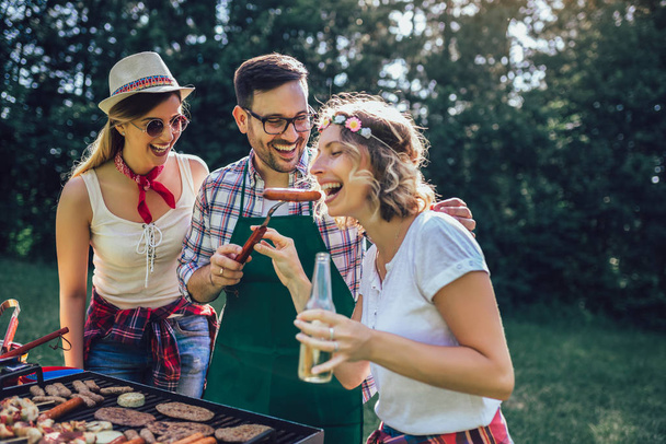 Group of friends stand at a barbecue, one cooking at grill - Foto, Imagen