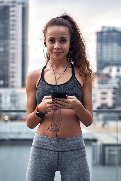 Young athletic woman in sport clothes standing on city background with phone in hands posting her training results - Fotografie, Obrázek