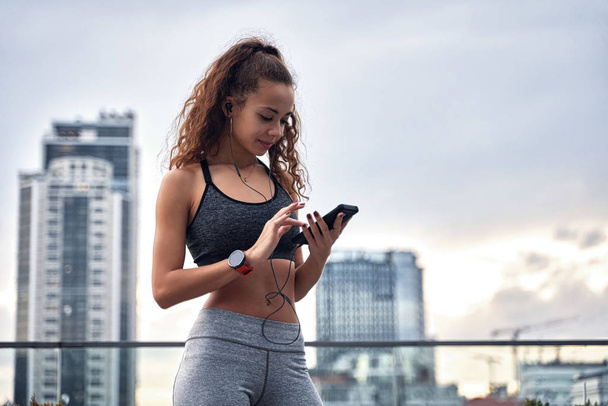 Молода спортсменка в спортивному одязі, стоячи на міському фоні з телефоном в руках, розміщуючи свої результати тренувань
 - Фото, зображення