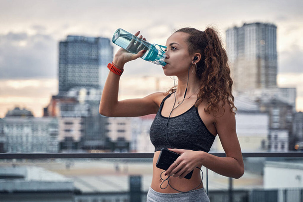Seitenaufnahme einer jungen athletischen Frau, die Wasser aus einer Sportflasche trinkt, während sie Musik vor dem Hintergrund der Stadt hört - Foto, Bild