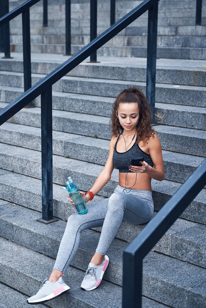 Young athletic woman in sport clothes sitting on stairs of city stadium with phone in hands posting her training results - Photo, Image