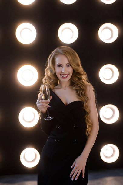 A modern festive young woman, blonde with curly long hair, in a black dress with French manicure. In the hands of a glass of champagne. On Background of bright led lamps. Fashion girl copy space   - Foto, imagen