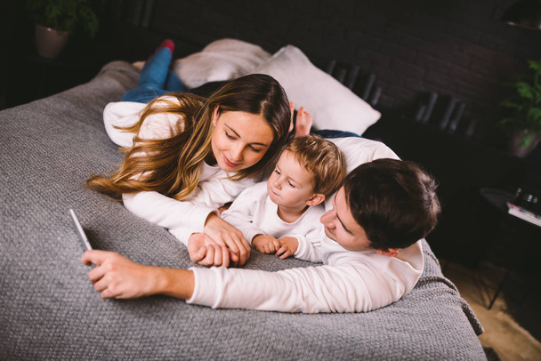 Μια νεαρή οικογένεια ξαπλώνει στο κρεβάτι και κοιτάζει το κινητό τηλέφωνο. Μαμά, ο μπαμπάς και ο γιος βλέπουν ένα βίντεο σε ένα smartphone στην κρεβατοκάμαρα το βράδυ. Οικογενειακές απογευματινές διακοπές στο σπίτι - Φωτογραφία, εικόνα