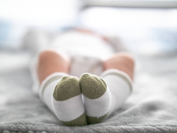 Μια κοντινή φωτογραφία ενός νεογέννητου αγοριού χαριτωμένο πράσινο αιχμή άσπρες κάλτσες όπως ο ίδιος ξαπλώνει σε μια γκρι κουβέρτα στον ήλιο με το σώμα θολή και από την εστίαση μακριά από την κάμερα και τα πόδια κοντά στην κάμερα. - Φωτογραφία, εικόνα