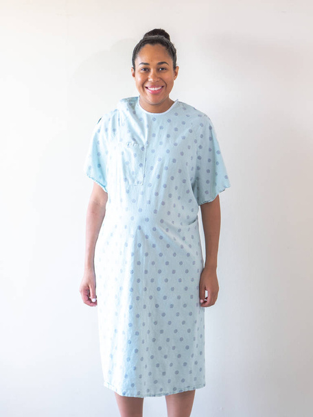 A beautiful mixed race African American woman with her hair put up in a bun looks at the camera and smiles big wearing a blue patterned hospital gown or robe as she receives good news from her doctor. - Photo, Image
