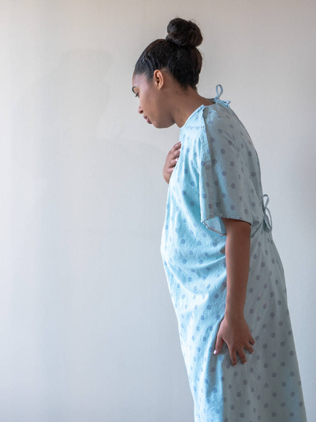 Uma jovem de raça mista afro-americana vestindo um vestido de hospital ou roupão olha para baixo e se inclina enquanto segura o peito com a mão com um olhar de desconforto náusea ou tristeza
. - Foto, Imagem