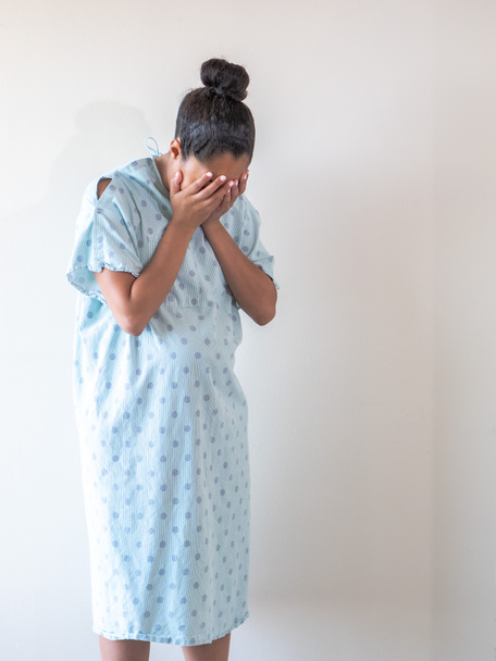 Uma jovem mulher negra americana de raça mista grávida vestindo um vestido de hospital ou roupão coloca ou esconde seu rosto em suas palmas abertas enquanto ela palpita lágrimas de tristeza ou depressão
. - Foto, Imagem
