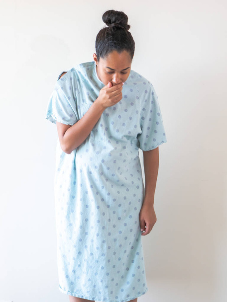A beautiful pregnant middle aged mixed race African American woman with her hair up in a bun wearing a hospital gown and sick or ill coughs into her hand and looks down at the ground. - Photo, Image