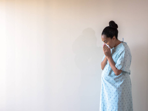 Молодая красивая афро-американская женщина смешанной национальности с обычной простудой или гриппом высморкается в белую бумагу для лица, когда чихает в узорчатом больничном платье
. - Фото, изображение