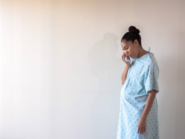 A young postpartum mixed race African American woman wearing a hospital gown or robe hunches over and looks down as she dries her eyes from tears of sadness or depression from a loss. - Foto, imagen