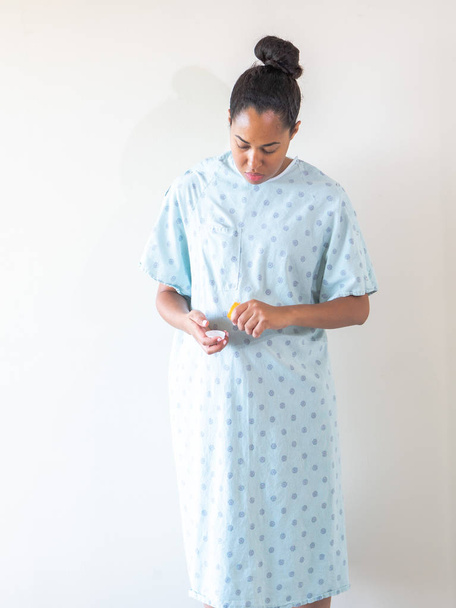 Uma jovem de raça mista afro-americana vestindo um vestido de hospital azul padronizado ou robe toma pílulas de medicação de um recipiente de plástico e derrama-los em sua mão para consumir
. - Foto, Imagem