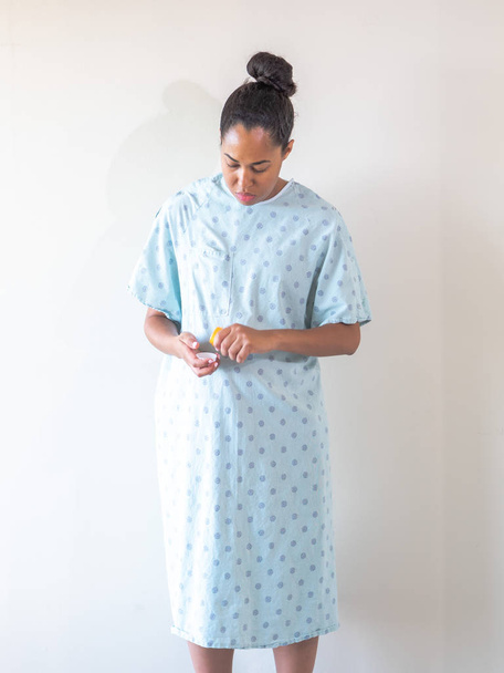 Een jonge gemengde ras Afro-Amerikaanse vrouw draagt een blauw patroon ziekenhuisjurk of badjas neemt medicatie pillen uit een plastic container en giet ze in haar hand om te consumeren. - Foto, afbeelding