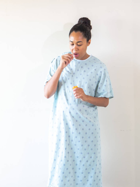 Een jonge gemengde ras Afro-Amerikaanse vrouw draagt een blauw patroon ziekenhuisjurk of badjas neemt medicatie pil uit een plastic container en zet ze in haar mond om te consumeren. - Foto, afbeelding