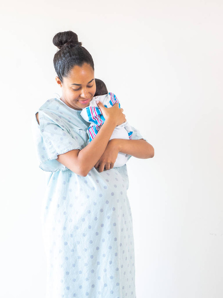 Мішана раса, афроамериканська мати, одягнена в лікарняну сукню, дивиться вниз на своє зовсім нове немовля, коли вона тримає його на плечі обіймаючись і колихаючи його, коли він спить в ковдрі болота. - Фото, зображення