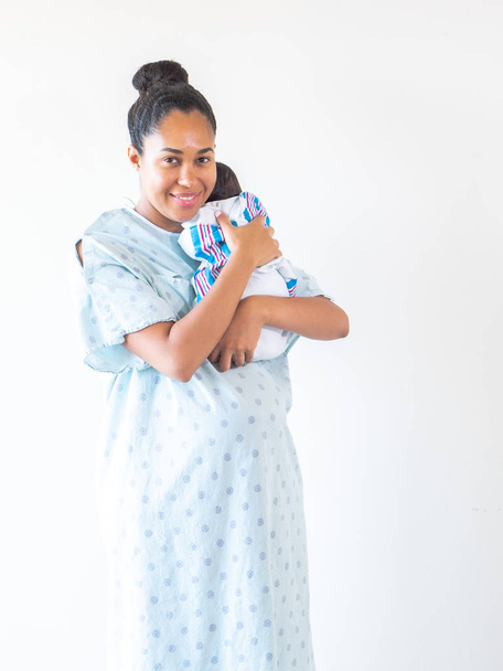混合レースアフリカ系アメリカ人の母親を着て病院のガウン彼は彼の毛布のスワドルで眠るように彼女の肩の抱擁に彼女のブランドの新しい幼児の赤ちゃんを保持し、彼を圧着. - 写真・画像
