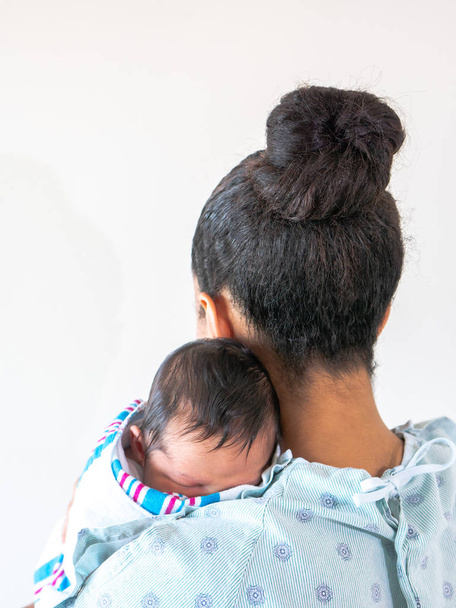Une mère afro-américaine de race mixte portant une robe d'hôpital tient son tout nouveau bébé garçon avec beaucoup de cheveux sur son épaule l'étreignant et le berçant alors qu'il dort dans sa lange de couverture
. - Photo, image