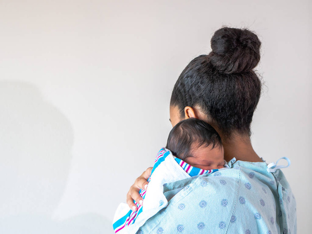 Een gemengd ras Afro-Amerikaanse moeder kijkt weg van de camera het dragen van een ziekenhuis toga houdt haar gloednieuwe baby over haar schouder knuffelen en wiegen hem als hij slaapt in zijn deken swaddle. - Foto, afbeelding