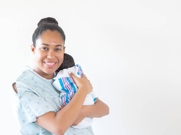 Een glimlachend gemengd ras Afro-Amerikaanse moeder draagt een ziekenhuisjurk houdt haar gloednieuwe baby op haar schouder knuffelen en wiegen hem als hij slaapt in zijn deken swaddle. - Foto, afbeelding