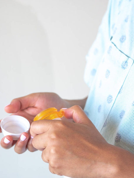 Een jonge gemengde ras Afro-Amerikaanse vrouw draagt een blauw patroon ziekenhuisjurk of badjas neemt medicatie pillen uit een plastic container en giet ze in haar hand om te consumeren. - Foto, afbeelding