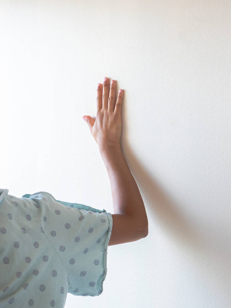 eine Nahaufnahme einer medizinischen Patientin im Krankenhauskleid, die ihren Arm ausstreckt und die Hand zur Unterstützung gegen die Wand hält, während sie Schmerzen oder Todesangst hat. - Foto, Bild