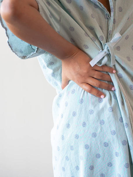 A close up photograph of a middle aged African American mixed race woman with her hand on her lower back or hip hunched over in pain wearing a patterned hospital gown. - Photo, Image