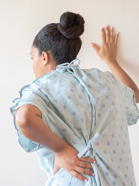 A close up photograph of a middle aged African American mixed race woman with her hand on her lower back or hip hunched over in pain and other hand on the wall wearing a patterned hospital gown. - Photo, Image