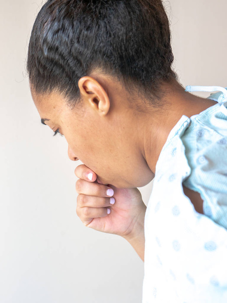 A close up side profile of a beautiful middle aged mixed race African American woman with her hair up in a hospital gown hunched over sick  or ill and coughing into her closed hand. - Photo, Image