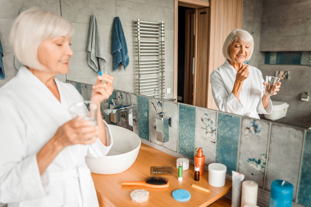 fröhliche alte Frau, die vor dem Spiegel steht und Tabletten nimmt - Foto, Bild