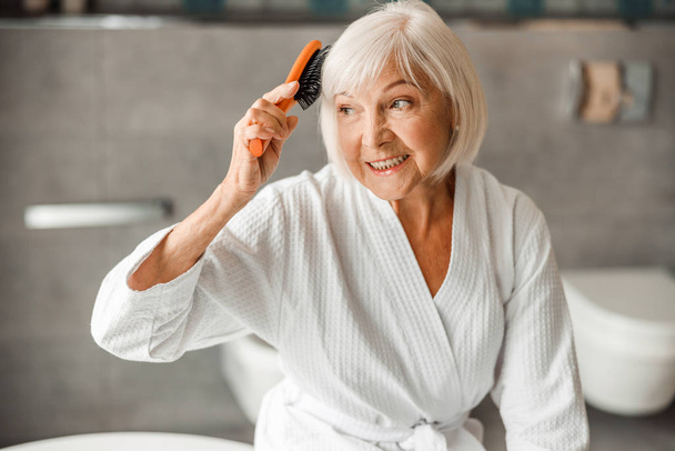 Preciosa anciana cepillando el cabello y sonriendo
 - Foto, imagen
