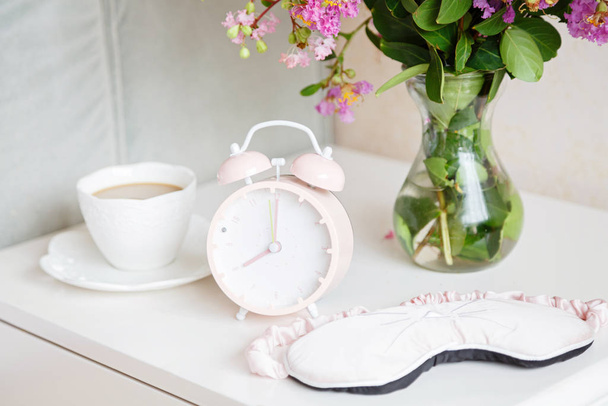 Alarm clock closeup, a cup of coffee and bouquet of pink flowers background in the morning sunlight - Fotografie, Obrázek
