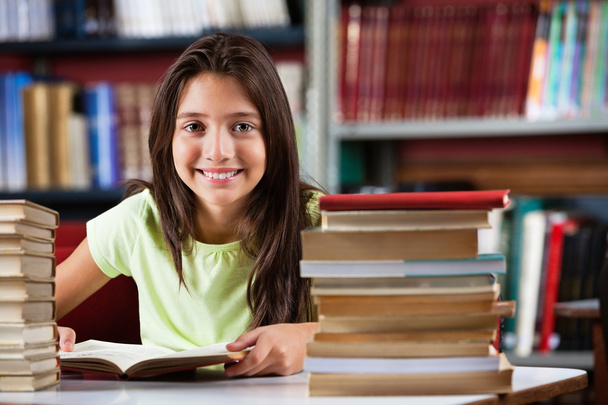 Schoolgirl Smiling While Sitting With Stack Of Books In Library - Foto, Bild