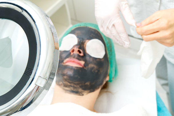 Young woman in a beauty salon with a black mask on her face. The concept of cosmetology. The cosmetologist performs a procedure to cleanse the face with problem skin. under cosmetic magnifying glass. - Photo, Image