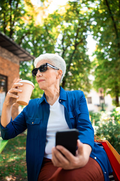 Senior woman drinking coffee and using cellphone outdoors - Foto, Bild
