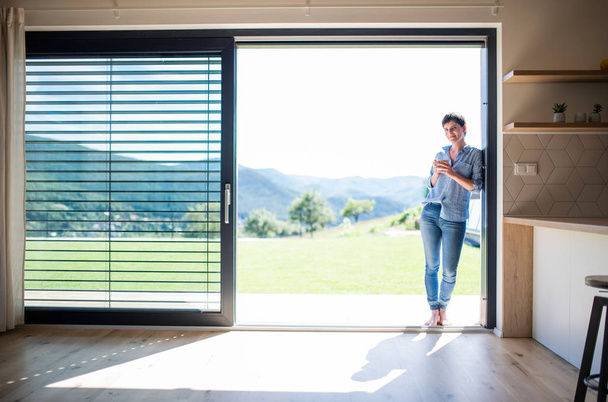 Front view of young woman with coffee standing by patio door at home. - Photo, Image