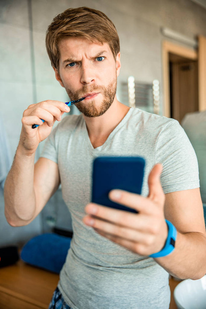 Bearded young man brushing teeth and holding cellphone - 写真・画像