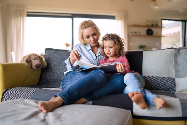 A cute small girl with mother on sofa indoors at home, reading book. - Foto, Imagem
