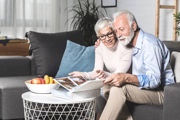 Romántica pareja de ancianos riendo mientras están sentados en el sofá en casa y viendo viejas fotos juntos
 - Foto, Imagen