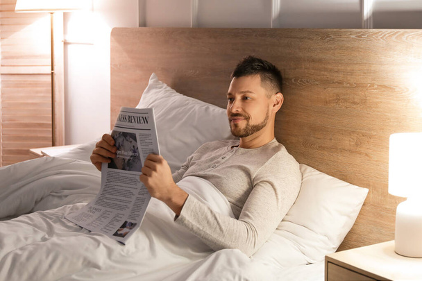 Όμορφος άντρας διαβάζει εφημερίδα ενώ είναι ξαπλωμένος στο κρεβάτι τη νύχτα. - Φωτογραφία, εικόνα