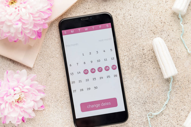 Kalendarz menstruacyjny na ekranie telefonu komórkowego i kobiecych produktów na szarym tle - Zdjęcie, obraz